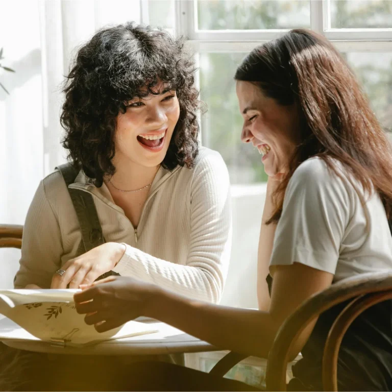 Two women laughing together at a table, engaging in conversation and sharing a moment of joy, emphasizing connection and support in a healing environment.