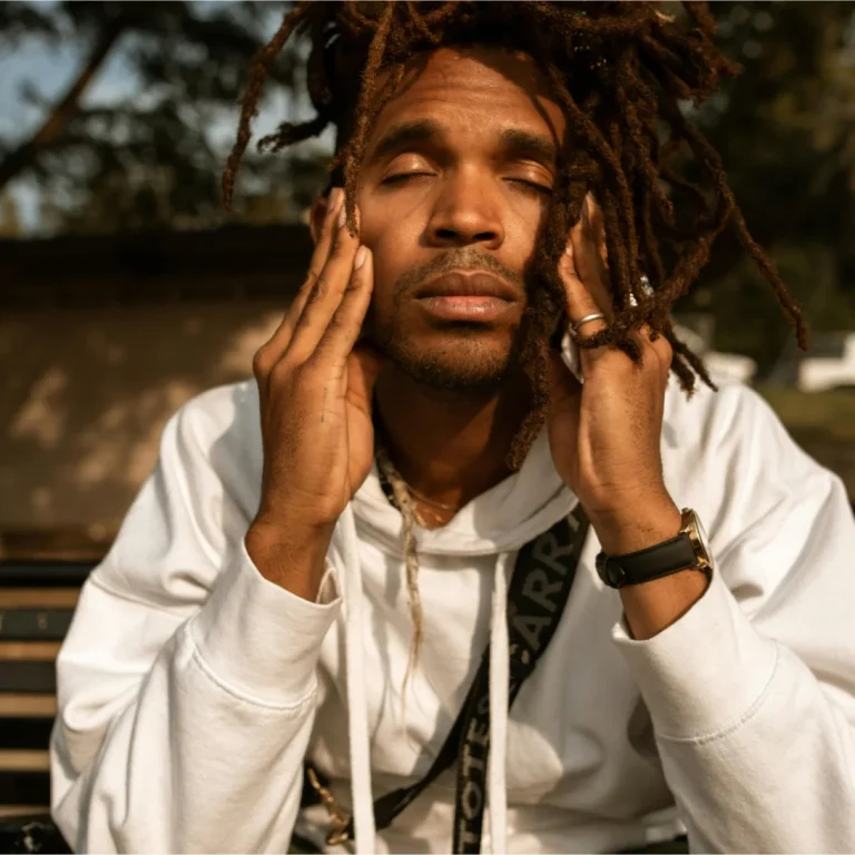 Man with dreadlocks wearing a white hoodie, eyes closed, hands on face, expressing a moment of contemplation or stress, symbolizing recovery and healing.